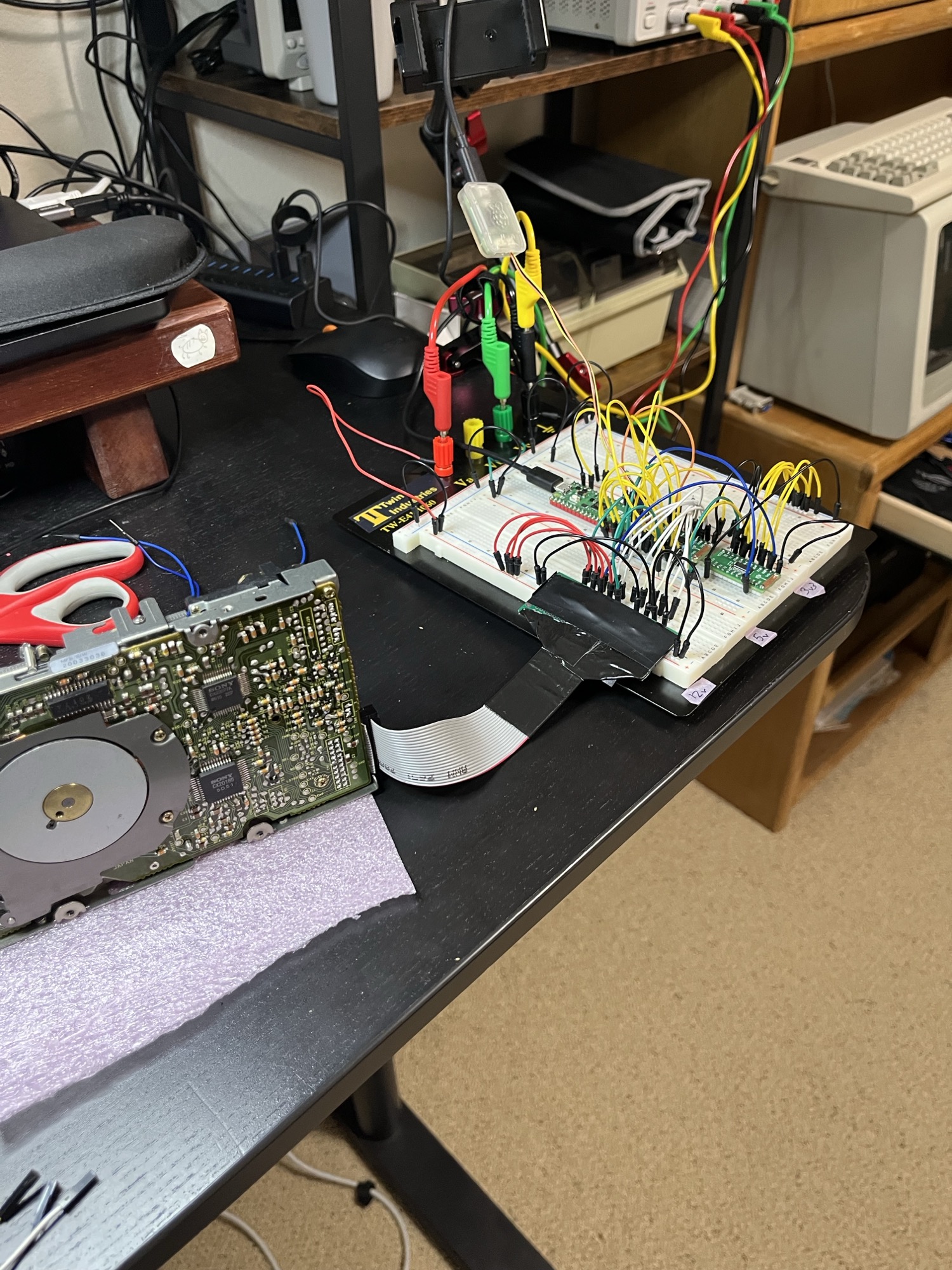 The M0 rig. A dead UniDisk's mechanism on the left, ribbon into the breadboard with the Pico 2 and two 8T245 boards, bench supply leads coming in from the shelf.